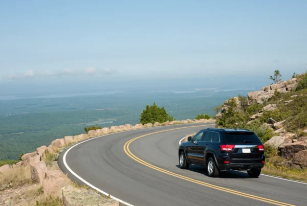 A black SUV driving around a curve on a two-lane mountain road above a forested valley and distant coastline on a clear summer day.