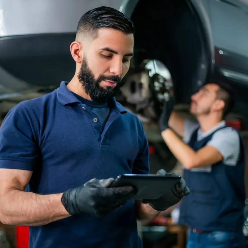 A mechanic in a navy polo and black gloves looks at a tablet while another technician works beneath a car raised on a lift with a wheel removed.