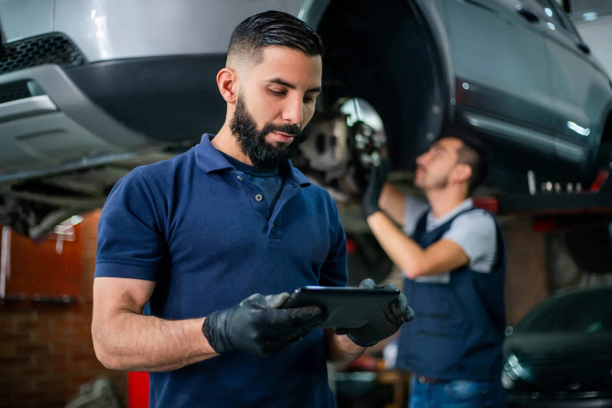 A mechanic in a navy polo and black gloves looks at a tablet while another technician works beneath a car raised on a lift with a wheel removed.