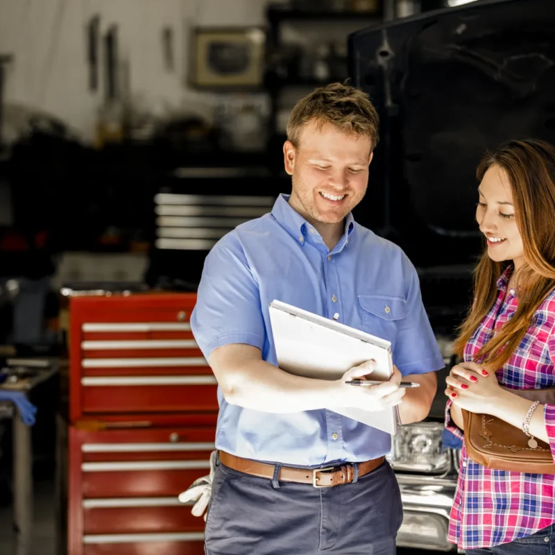 A smiling auto shop service advisor holding a clipboard reviews it with a female customer inside a repair bay with a red tool chest and a car with its hood open.