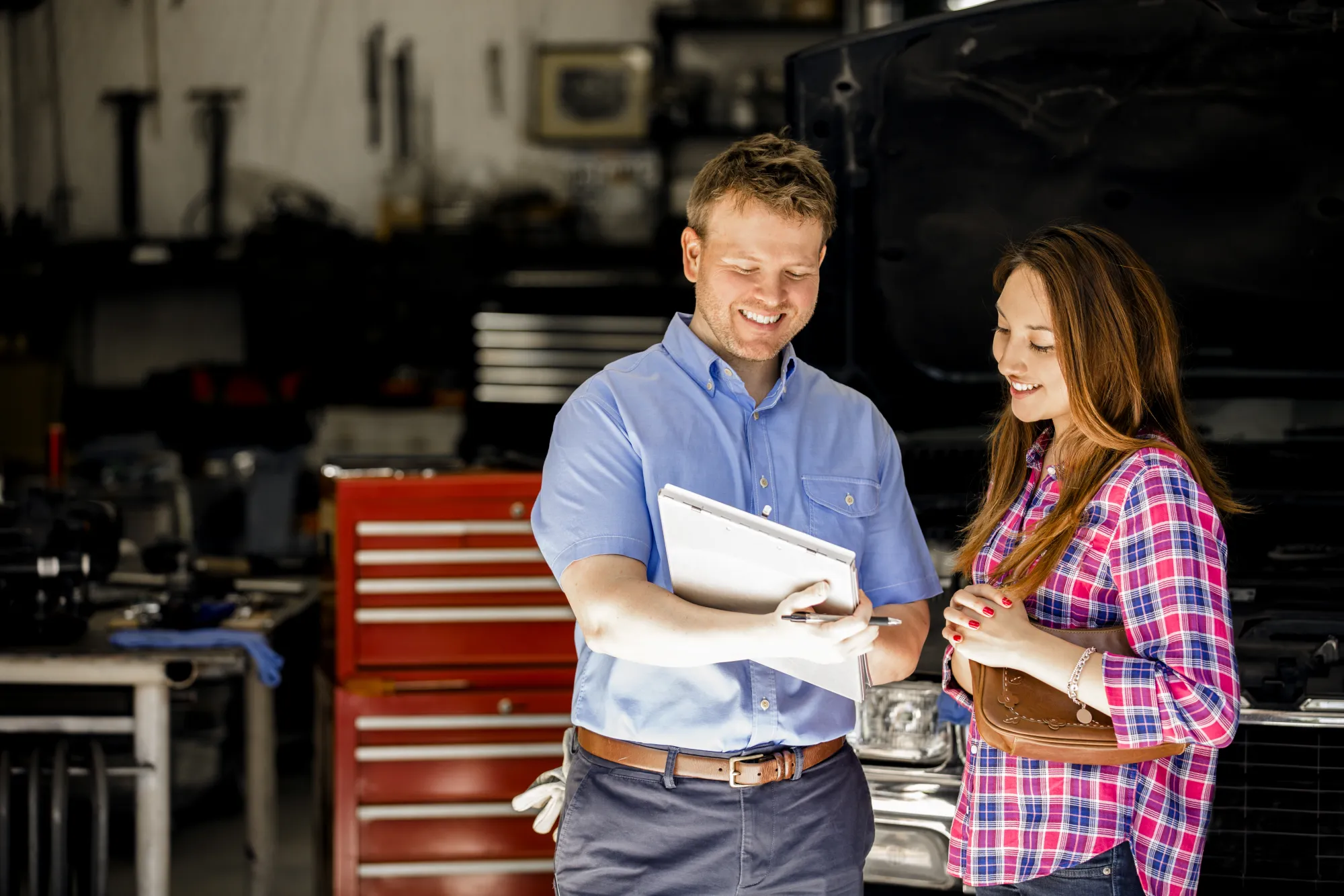 A smiling auto shop service advisor holding a clipboard reviews it with a female customer inside a repair bay with a red tool chest and a car with its hood open.